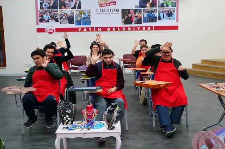 Kırmızı önlüklü atölye katılımcıları gülümseyerek ellerini havaya kaldırmış, kameraya selam veriyor.